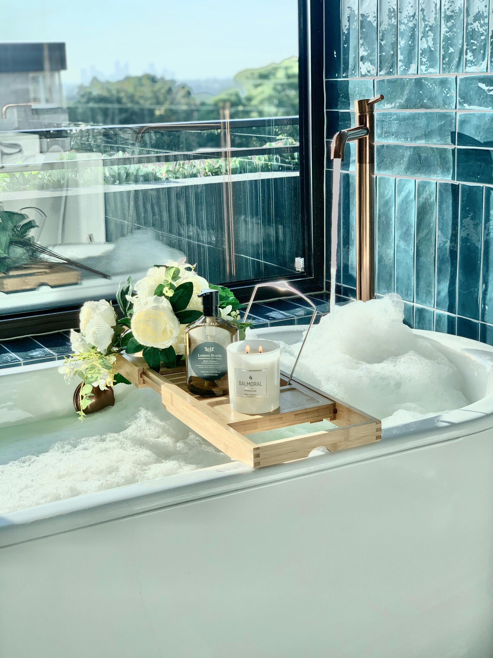 A spa-like bathtub setup with foam, a glowing candle, and fresh white roses on a wooden bath caddy.